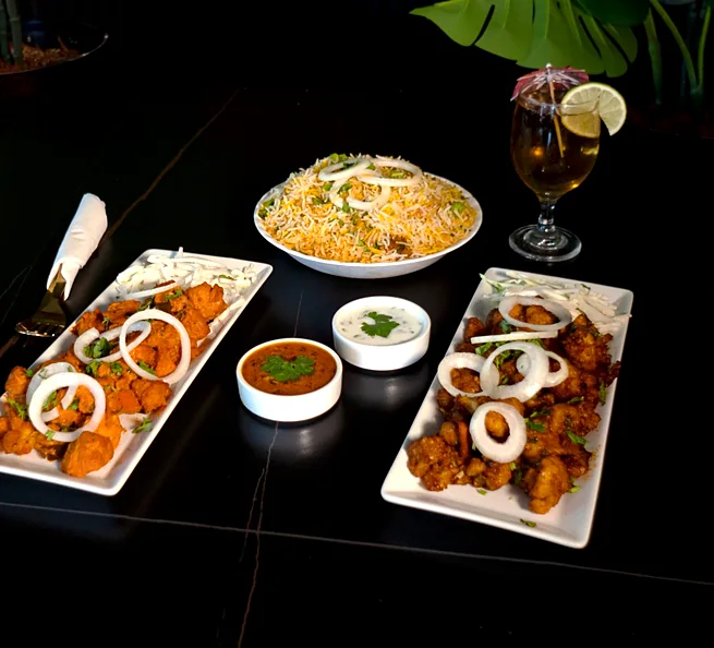Two platters of Chicken 65, a bowl of biryani, raita, salan and a drink on a dark table